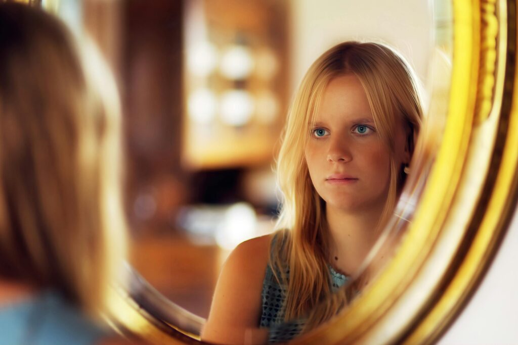 Vrouw kijkt met gemengde gevoelens naar zichzelf in de spiegel, twijfelend of ze moet toegeven aan wat haar ex wil.