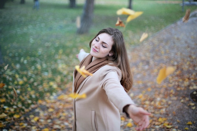 Jonge vrouw met gespreide armen en gesloten ogen, genietend van vallende herfstblaadjes, symboliseert loslaten en persoonlijke groei.