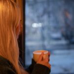 Jonge vrouw met kop koffie kijkt uit het raam en denkt na hoe ze haar ex terugkrijgt zonder spelletjes of manipulatie.