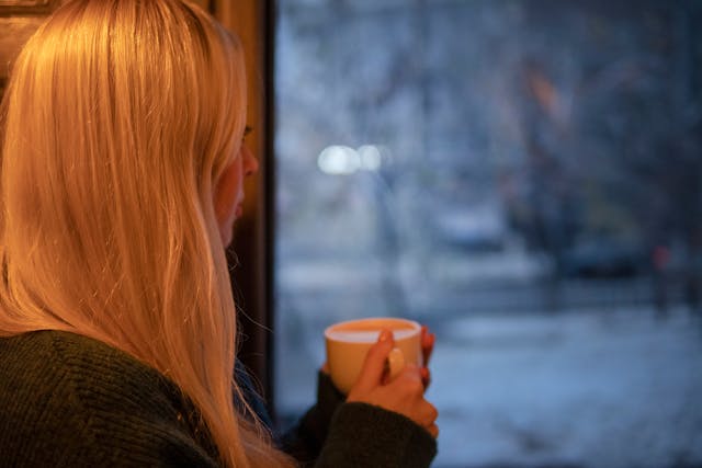 Jonge vrouw met kop koffie kijkt uit het raam en denkt na hoe ze haar ex terugkrijgt zonder spelletjes of manipulatie.